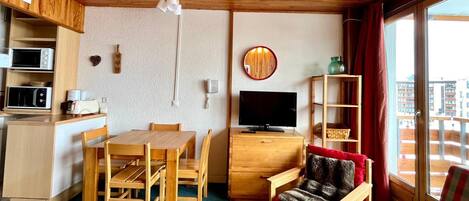A compact living space with a wooden ceiling, a dining table with chairs, a sofa, a television, and a small kitchen area with a microwave and toaster.