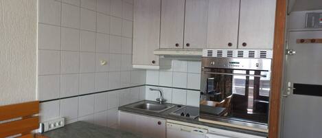 Een kleine keuken met witte tegels, houten keukenkastjes, een roestvrijstalen spoelbak en een inbouwoven.