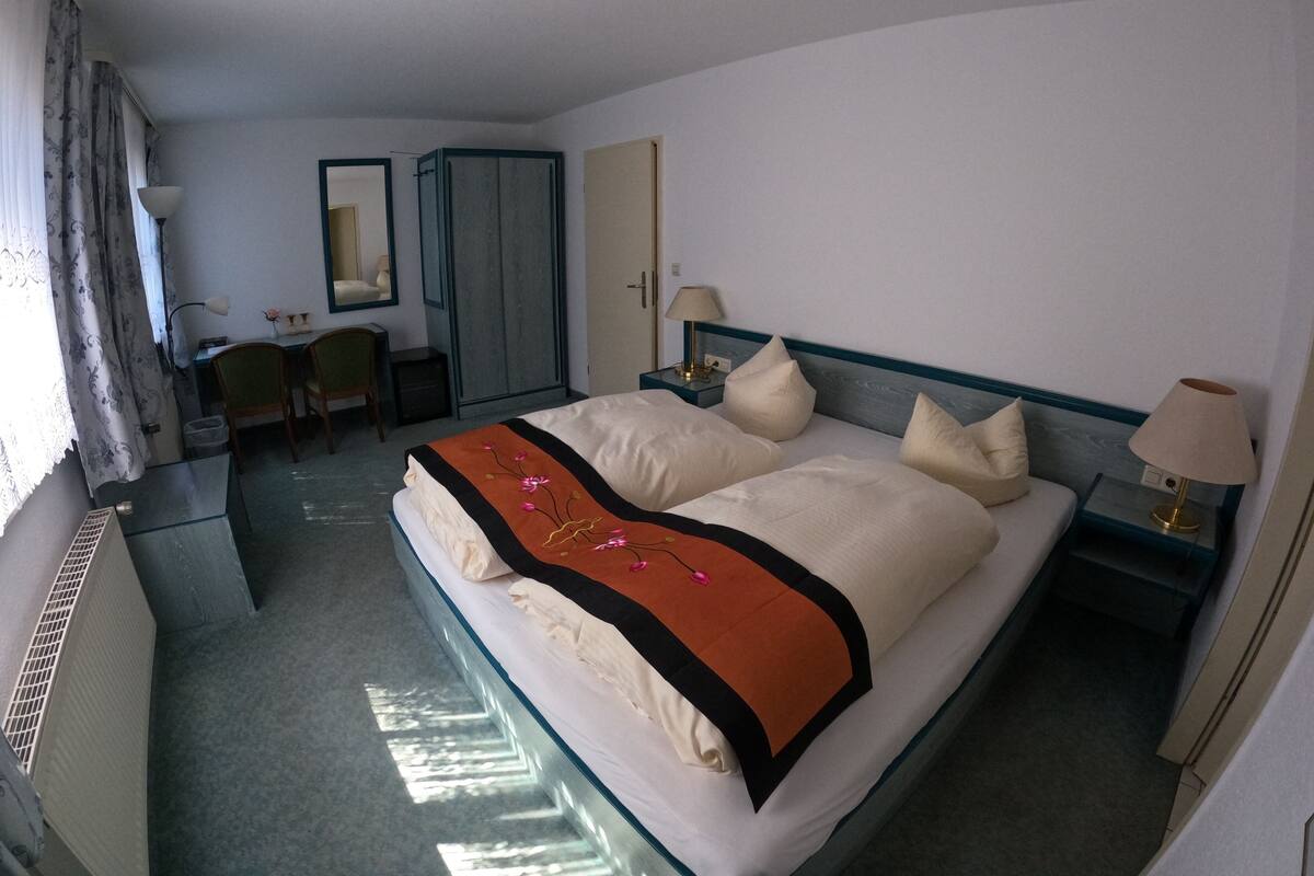 A hotel room with a large bed, a desk, a chair, a wardrobe, and a mirror.