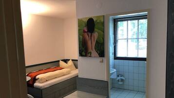 A small, compact room with a bed, a small table, and a bathroom with a window.