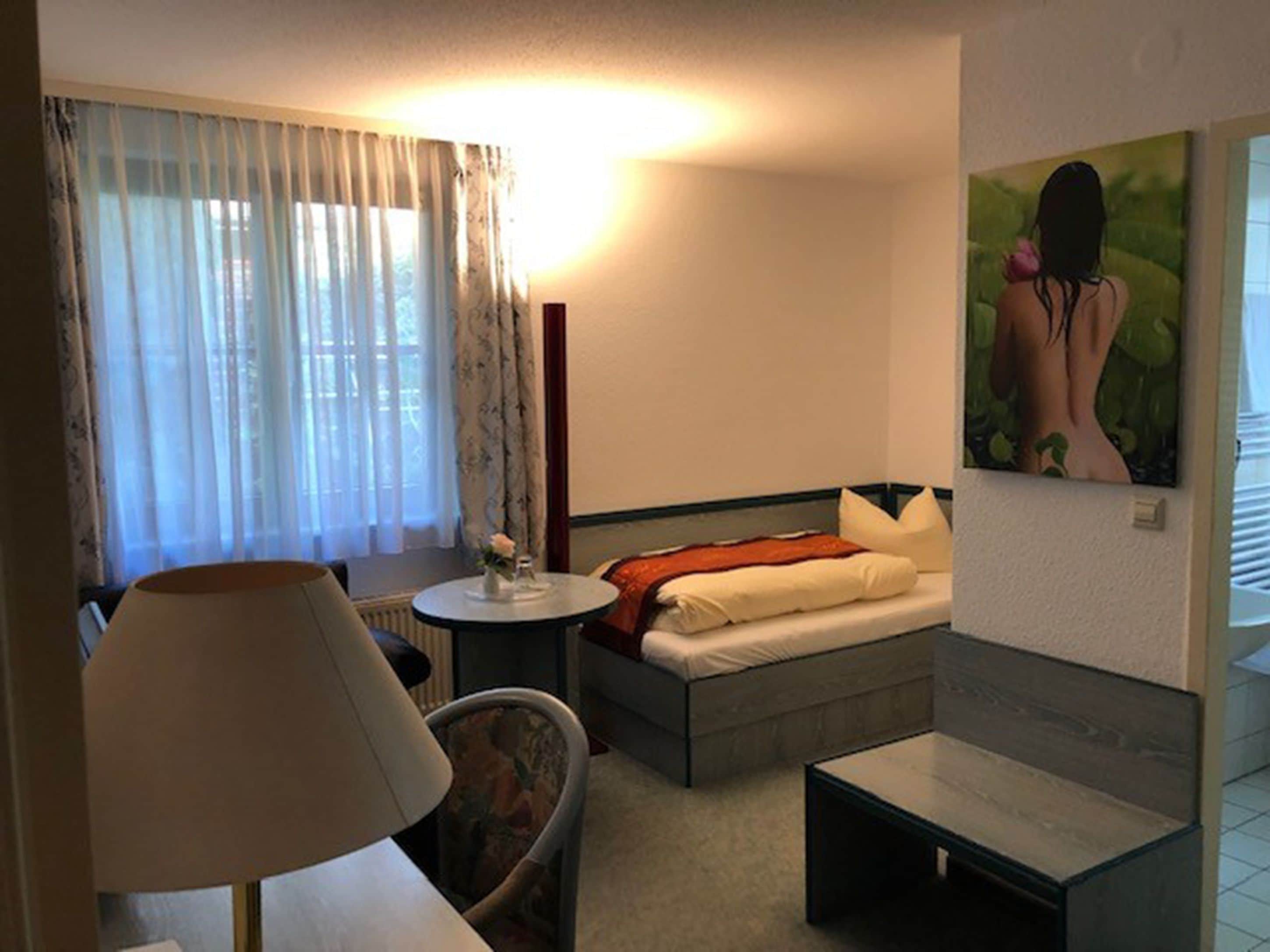A hotel room with a bed, a small round table, a chair, a lamp, and a painting on the wall.