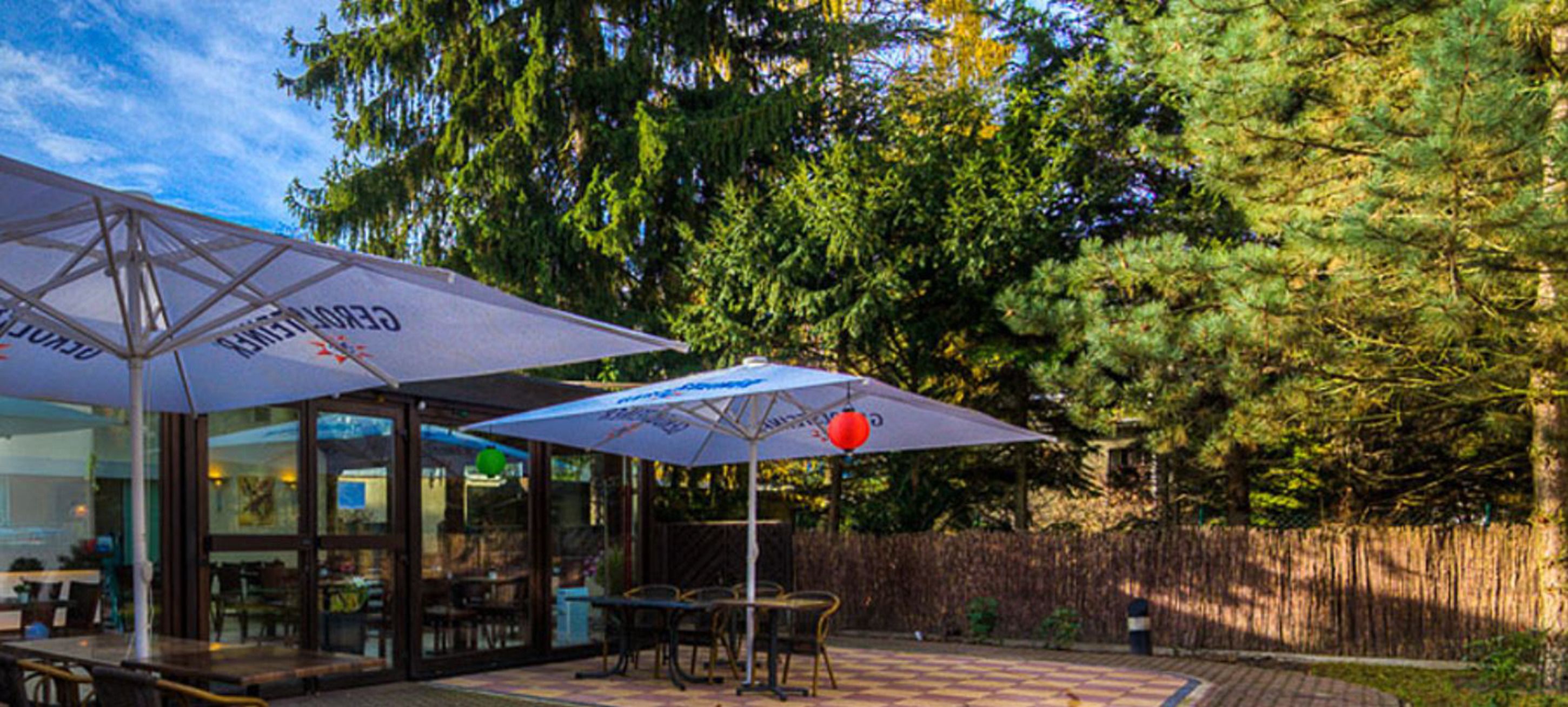 Een terras buiten met tafels en stoelen onder parasols, omgeven door een houten hek en bomen.