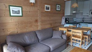 A cozy living room with a grey sofa, wooden dining table, and chairs. A kitchen with white cabinets and a hanging light fixture is visible in the background. - Studio with balcony and wifi 500m from slopes (Tignes)