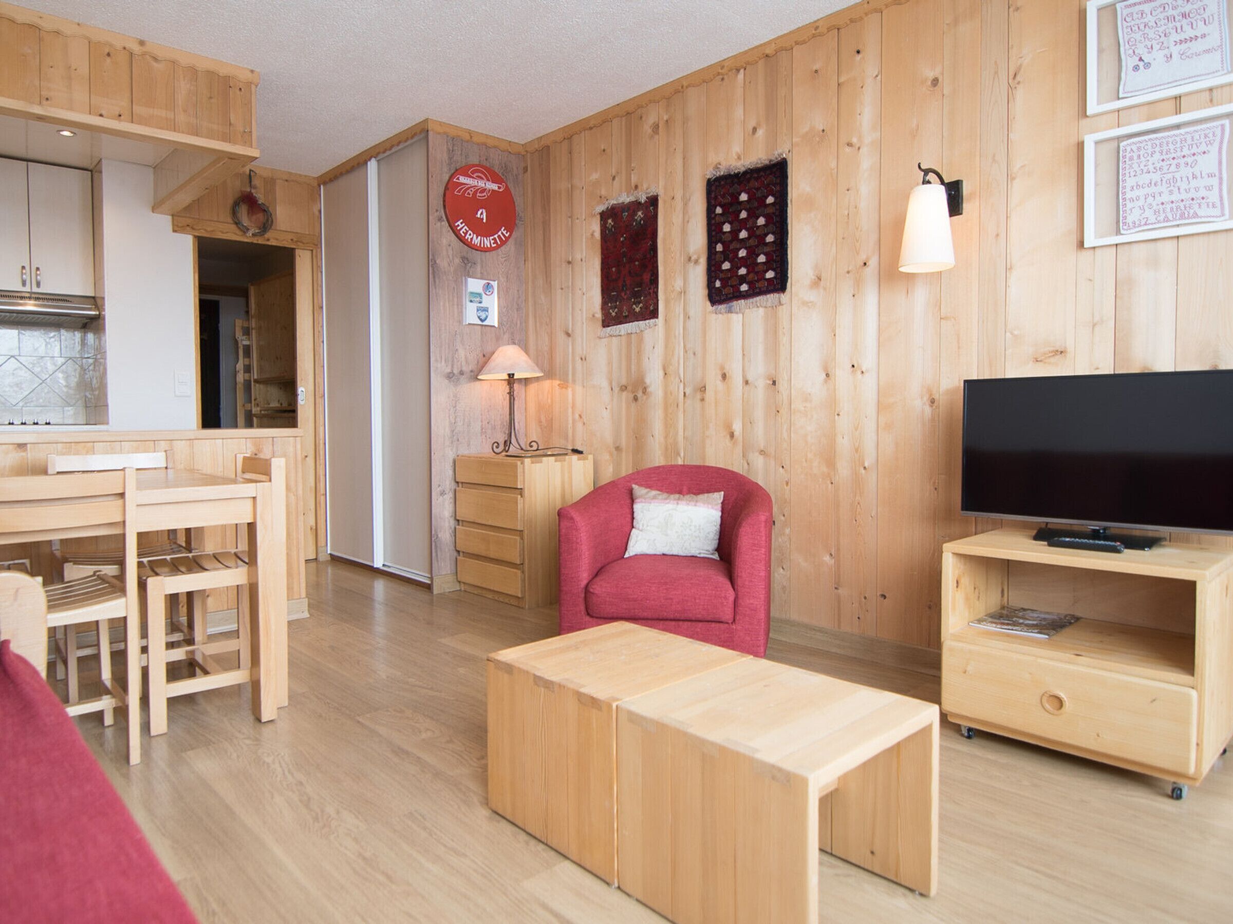A wooden-paneled room with a dining table, a red armchair, a TV on a wooden stand, and a lamp.