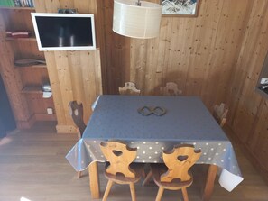 Una habitació amb panells de fusta, una taula coberta amb un mantell de punts, dues cadires de fusta amb motius de cor, un televisor penjat a la paret i una làmpada penjant.