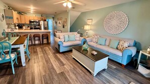A living room with a sofa, coffee table, and chairs. There is a kitchen area with a refrigerator, microwave, and stove.
