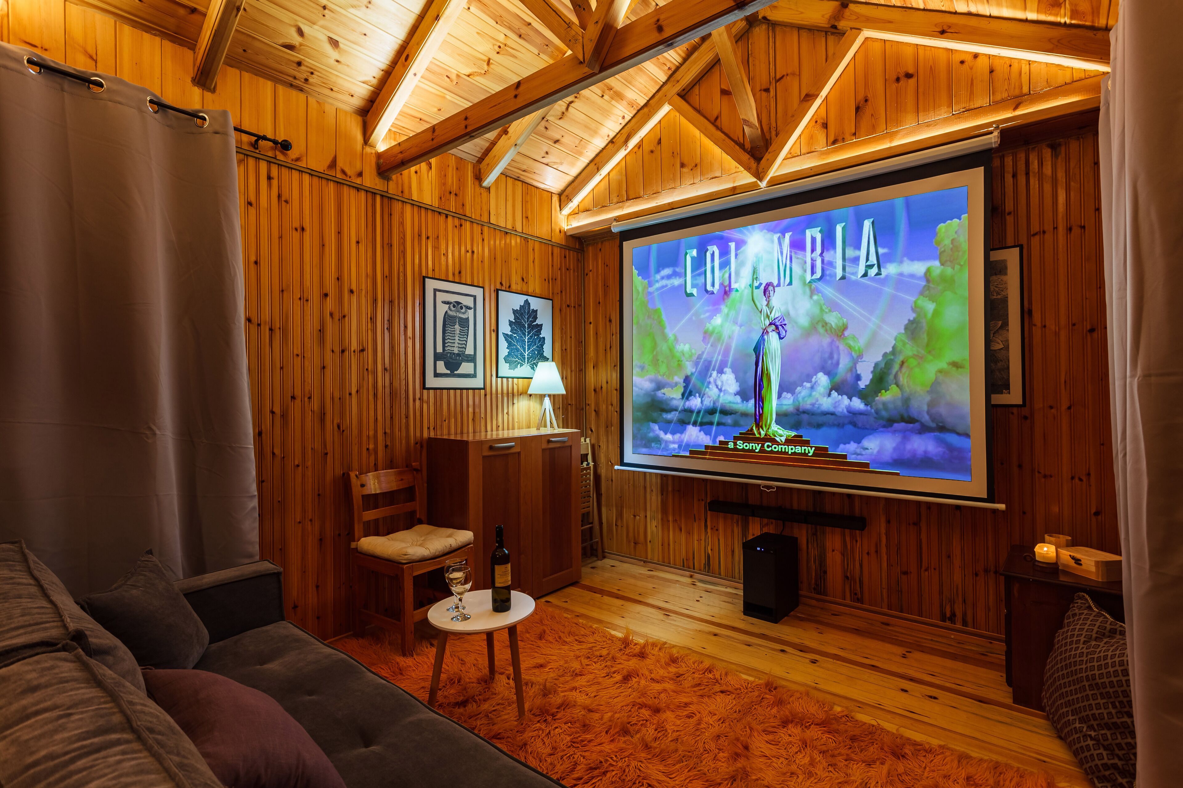 A cozy room with wooden walls, a large screen displaying 'Colombia', a sofa, a small table with a wine bottle, and a chair.