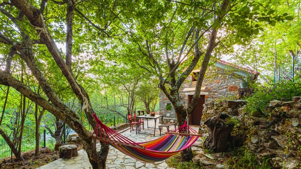 A hammock suspended between trees in a stone-paved area with a table and chairs, surrounded by greenery and a stone building. - Escape in the Forest, Cosy Chalet with Home Cinema (Kalamata)