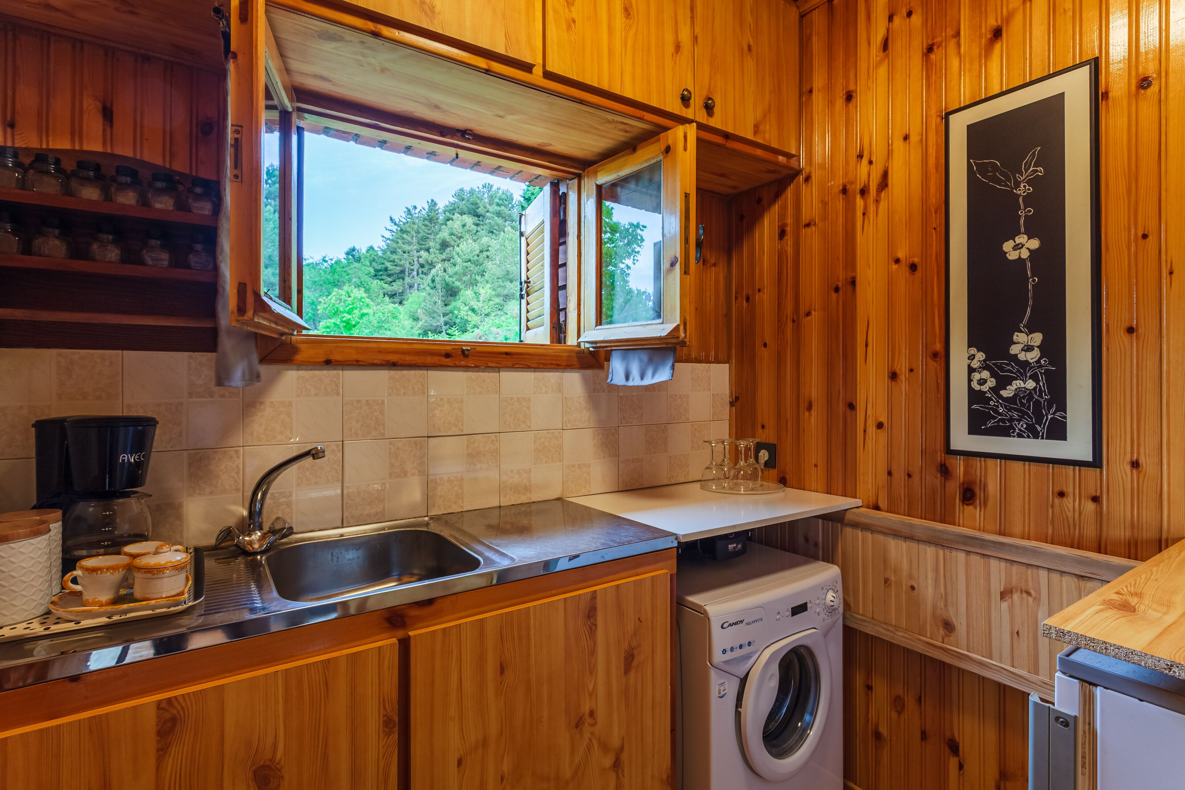 A wooden kitchen with a built-in washing machine, a sink, and a window with a view of trees.