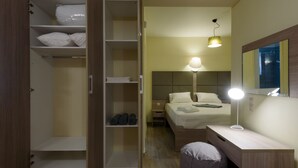 Una habitación de hotel con cama, escritorio con lámpara, espejo, armario y un pequeño cesto de basura.