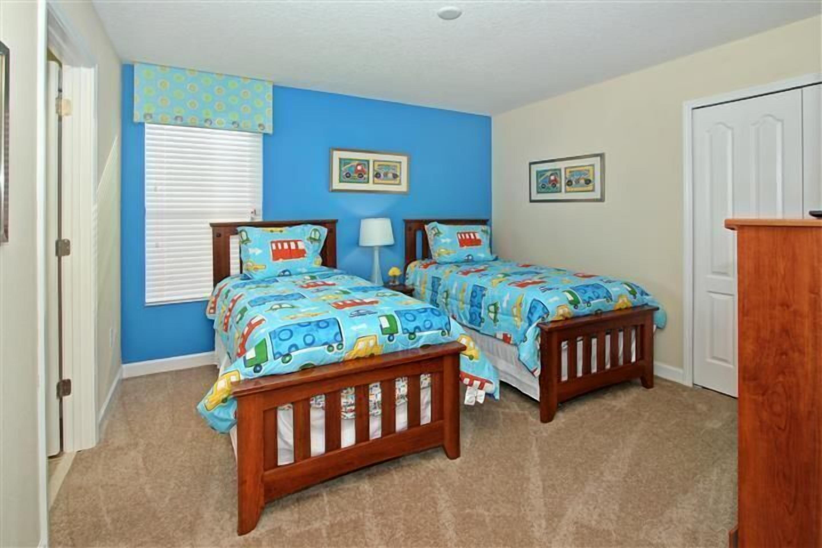 A bedroom with two beds, each with a blue and yellow train-themed bedspread, a wooden bench, and a window with blinds.