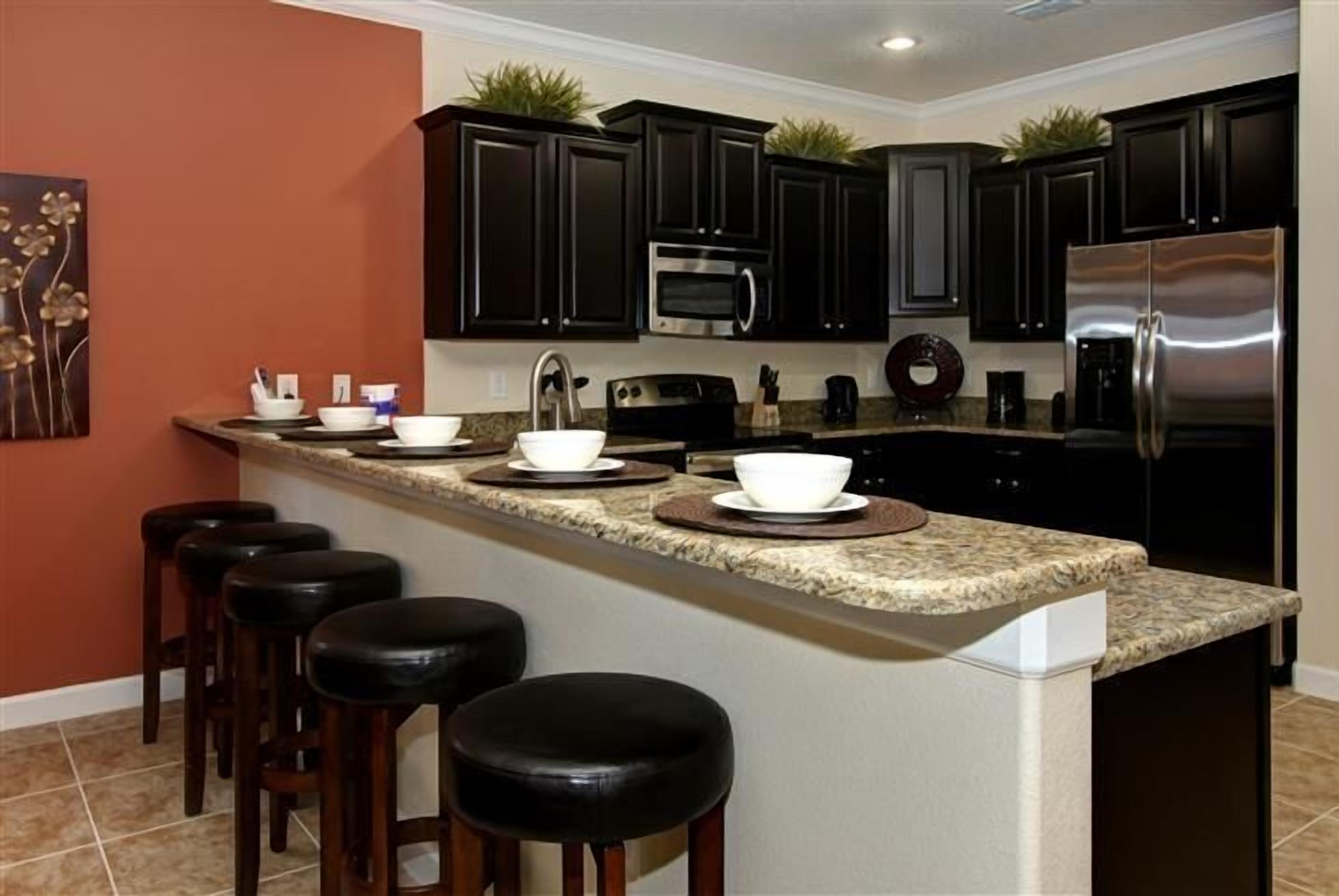 A modern kitchen with dark cabinets, a stainless steel refrigerator, a microwave, and a granite countertop. There are four stools and a decorative plant on the counter.