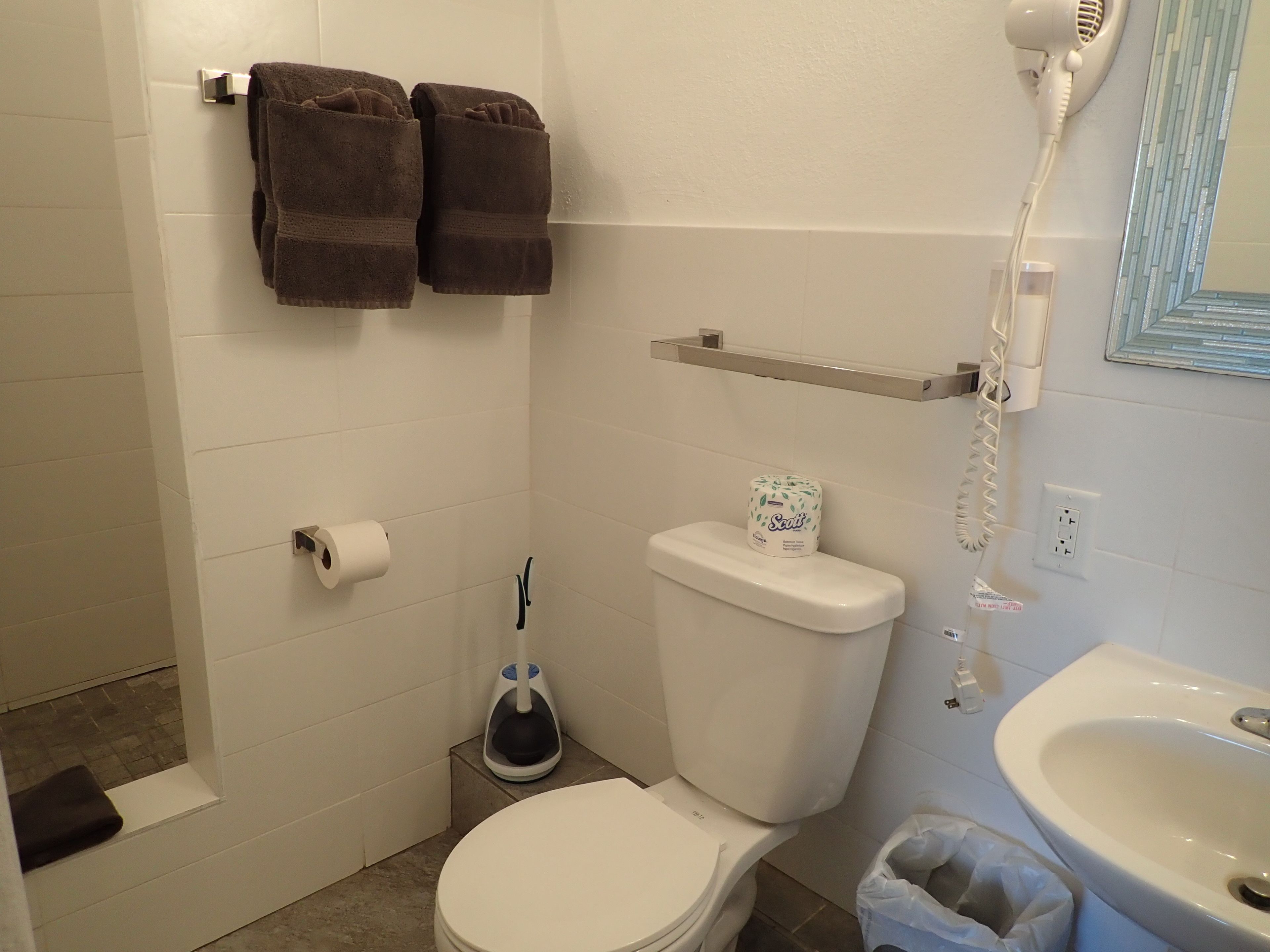 A bathroom with a toilet, a sink, a towel rack with towels, a toilet paper holder, a hairdryer, and a trash bin.
