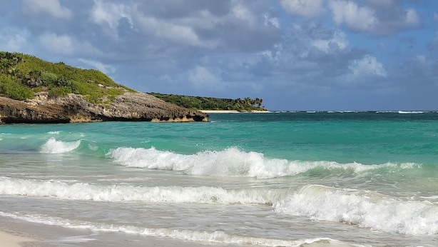 A beach with clear turquoise waters, waves crashing onto the shore, and a rocky cliff with vegetation in the background. - The Vieques Guesthouse (Vieques)