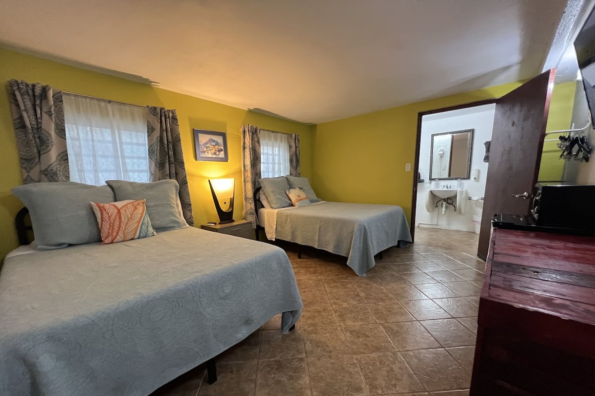 A room with two beds, a nightstand with a lamp, a bathroom visible through an open door, and a wooden cabinet.