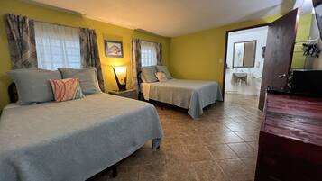 A room with two beds, a nightstand with a lamp, a bathroom visible through an open door, and a wooden cabinet.