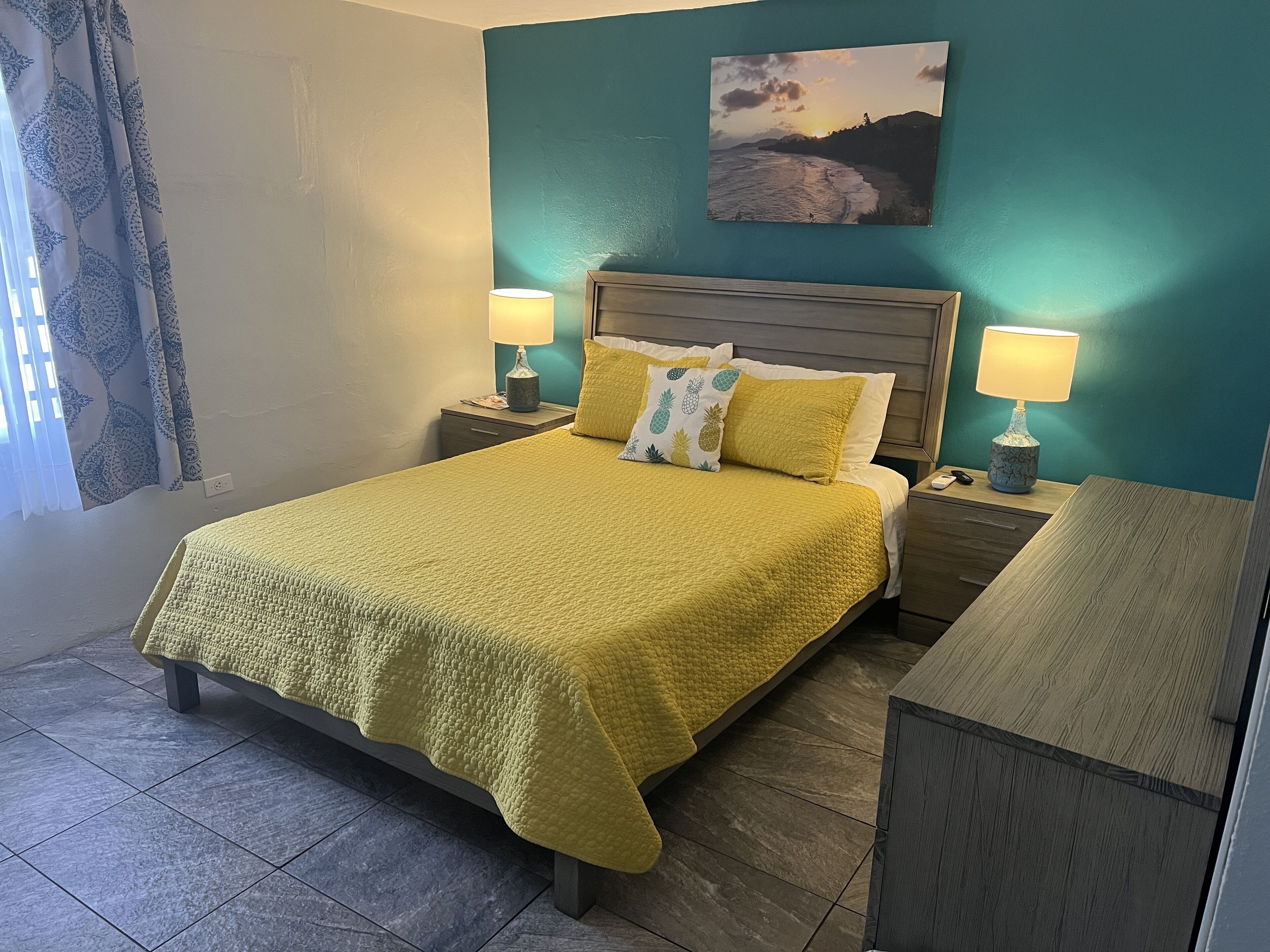 Une chambre d’hôtel bien rangée, avec un lit, deux tables de chevet dotées de lampes, un tableau au mur et une fenêtre avec des rideaux.