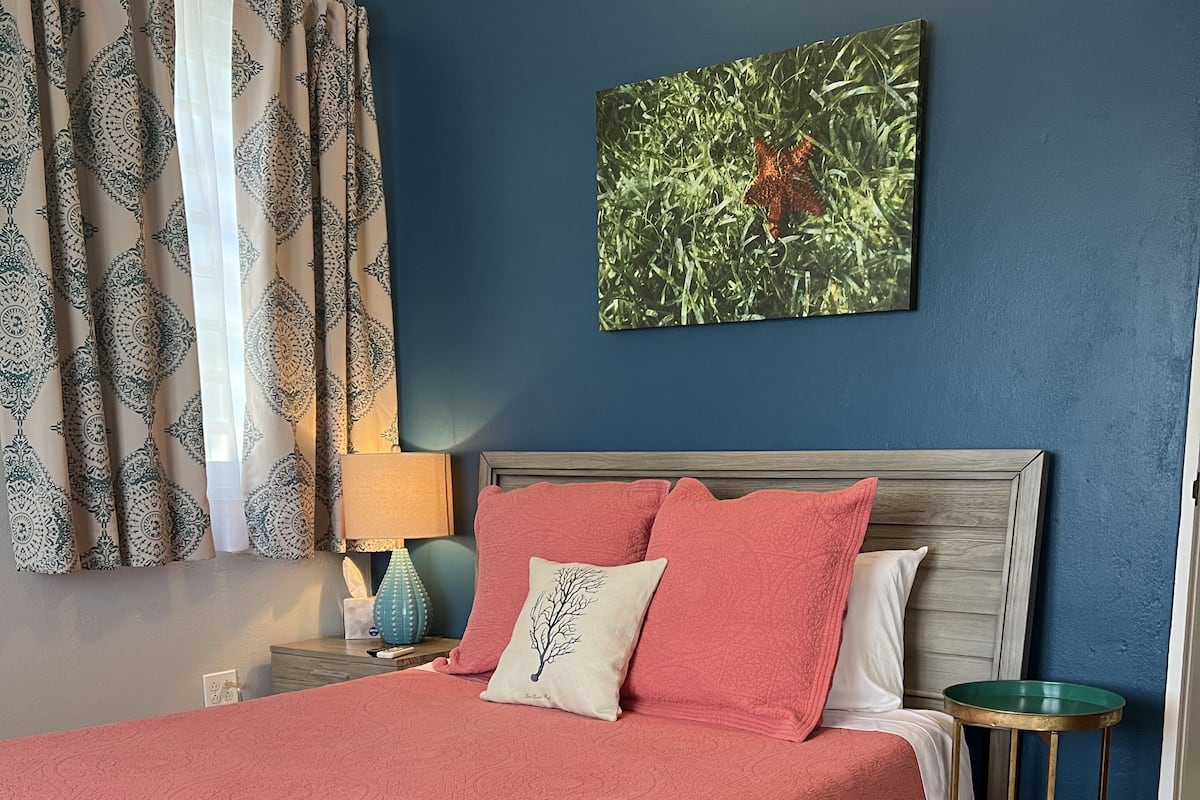 A bedroom with a bed, a nightstand, a lamp, a green side table, and a wall with a painting.