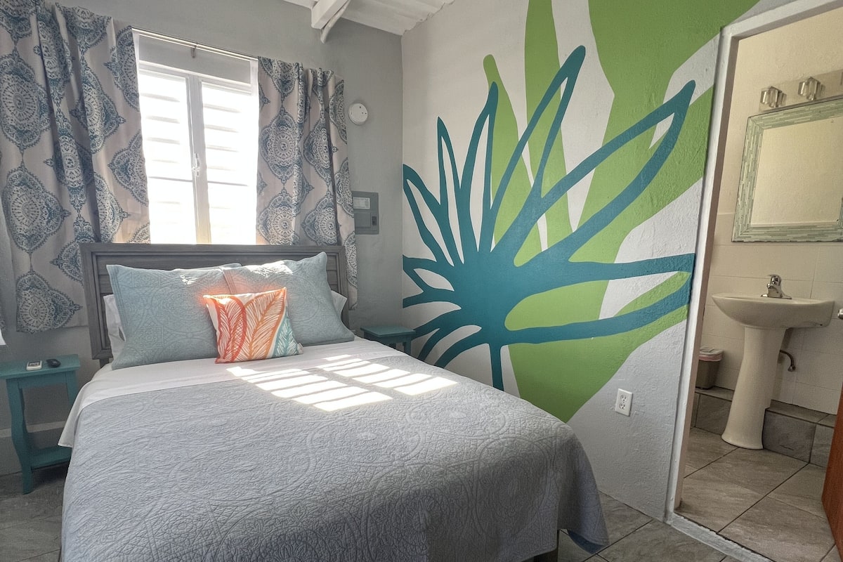 A bedroom with a bed, a window with patterned curtains, a green wall mural, and a bathroom with a sink and mirror.