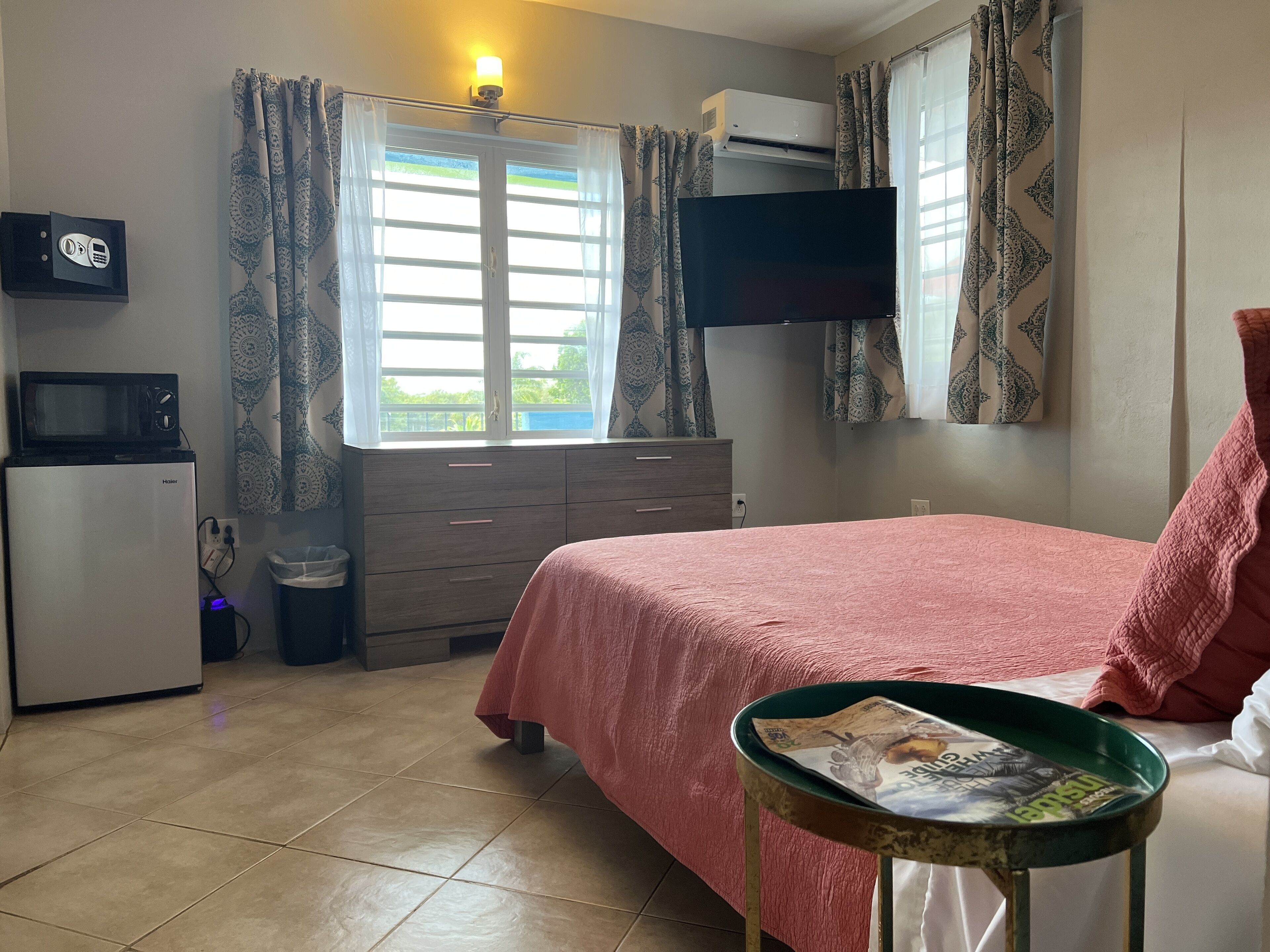 Un dormitorio con cama, una mesita con un periódico, televisor, aire acondicionado, microondas y refrigerador.
