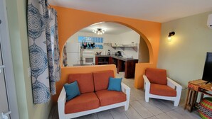 A living room with a sofa and armchair, an archway leading to a kitchen, and a television on a wooden stand. - The Vieques Guesthouse (Vieques)
