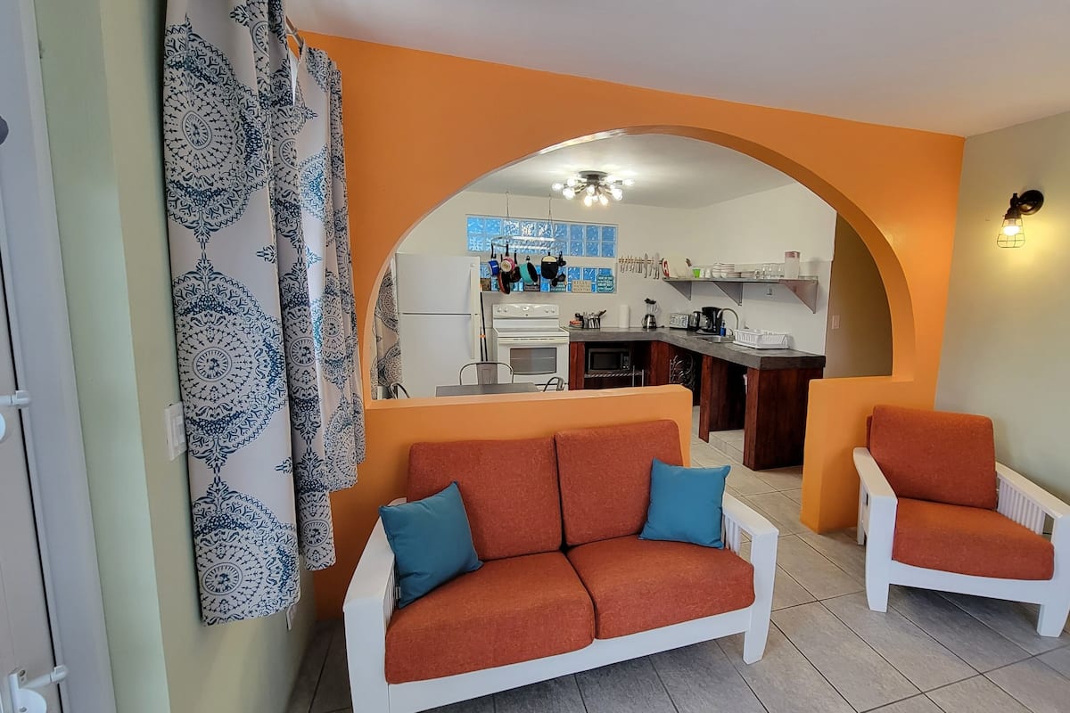 A living room with a sofa and armchair, an archway leading to a kitchen, and a television on a wooden stand.