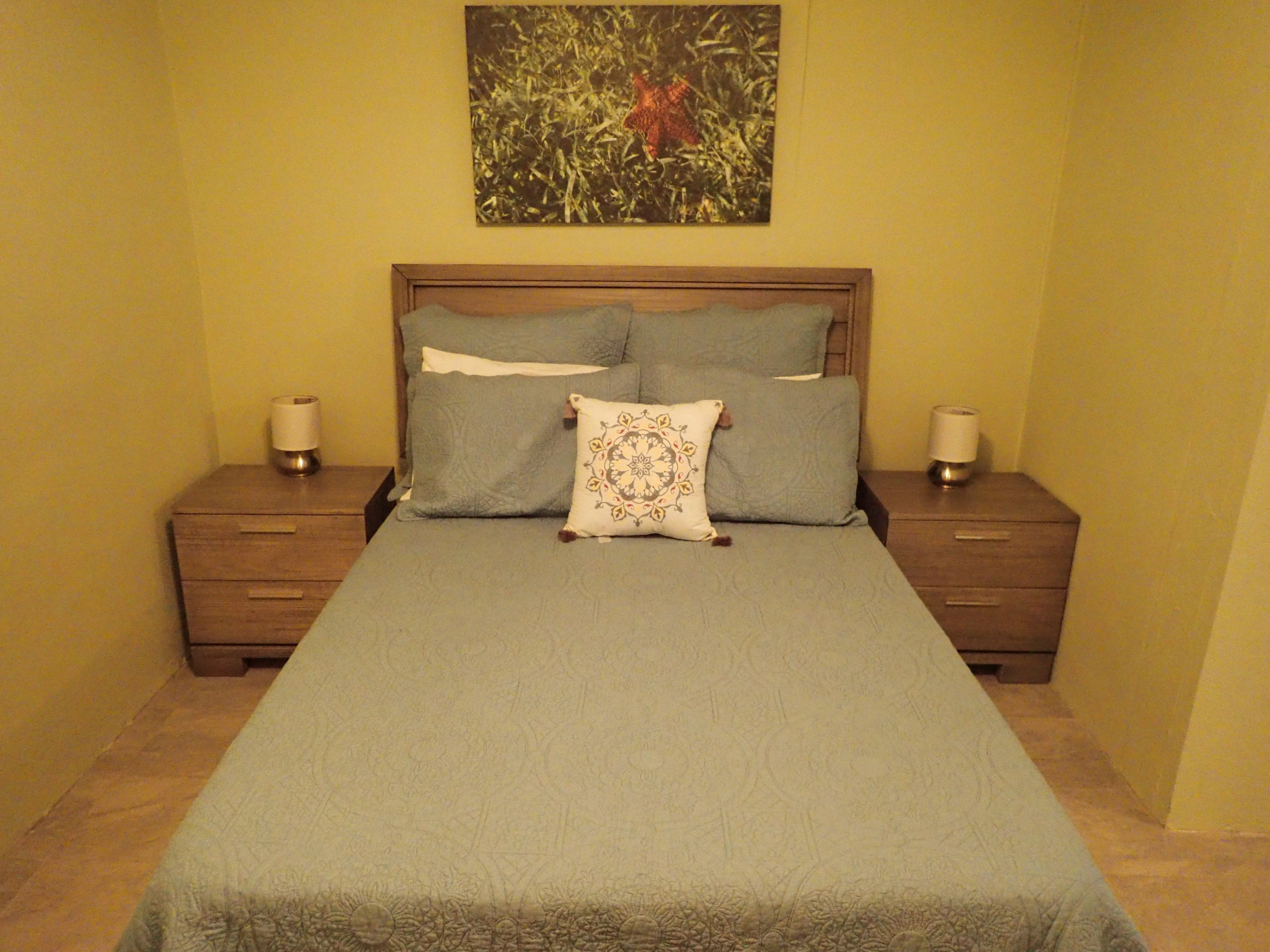 A neatly made bed with a blue bedspread and decorative pillows, flanked by wooden nightstands with lamps, in a room with yellow walls and a painting above the bed.