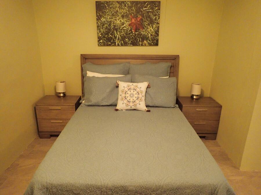 A neatly made bed with a blue bedspread and decorative pillows, flanked by wooden nightstands with lamps, in a room with yellow walls and a painting above the bed.