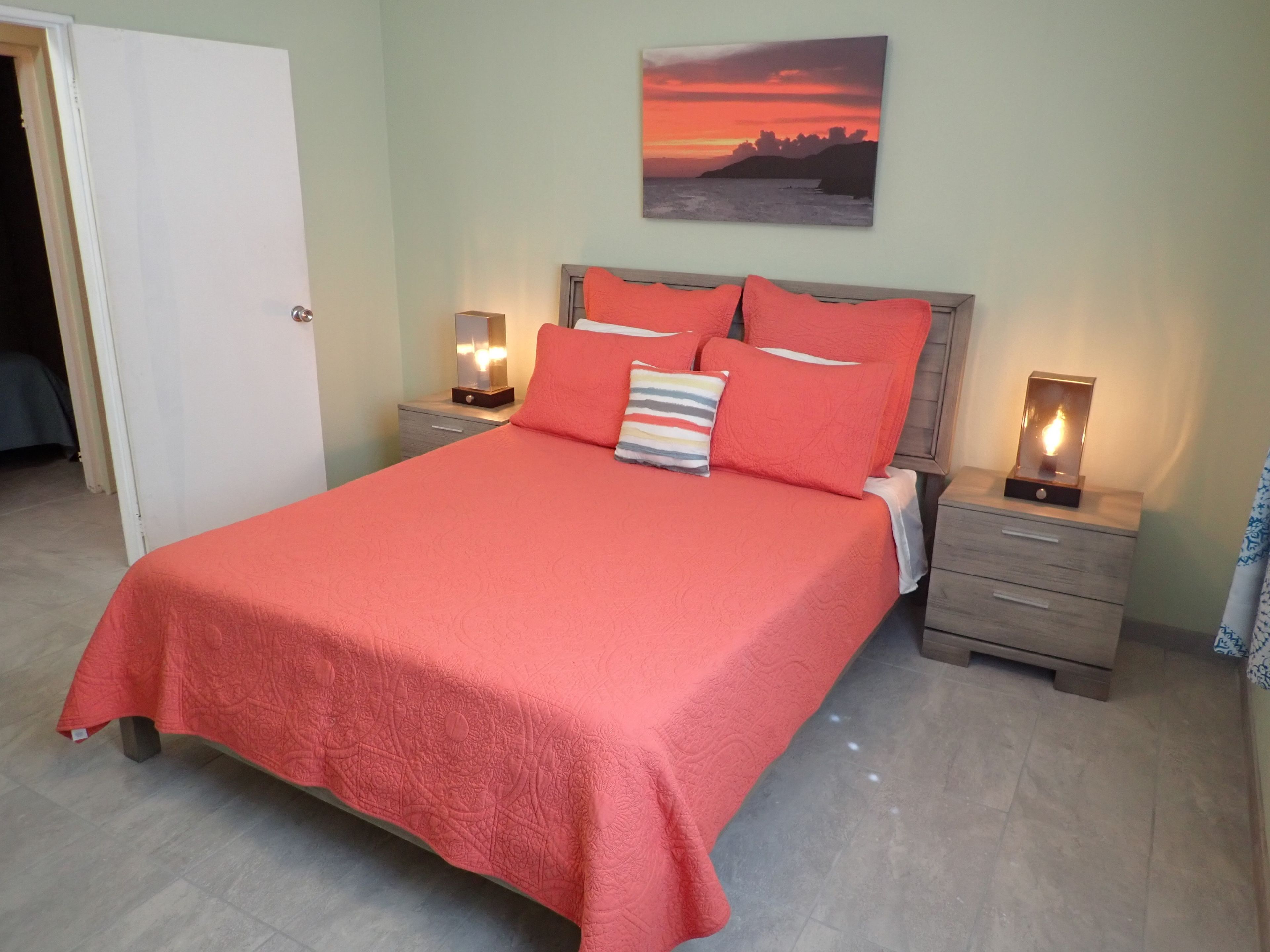 A neatly made bed with a red bedspread, two bedside tables with lamps, a painting of a sunset on the wall, and a door to the left.
