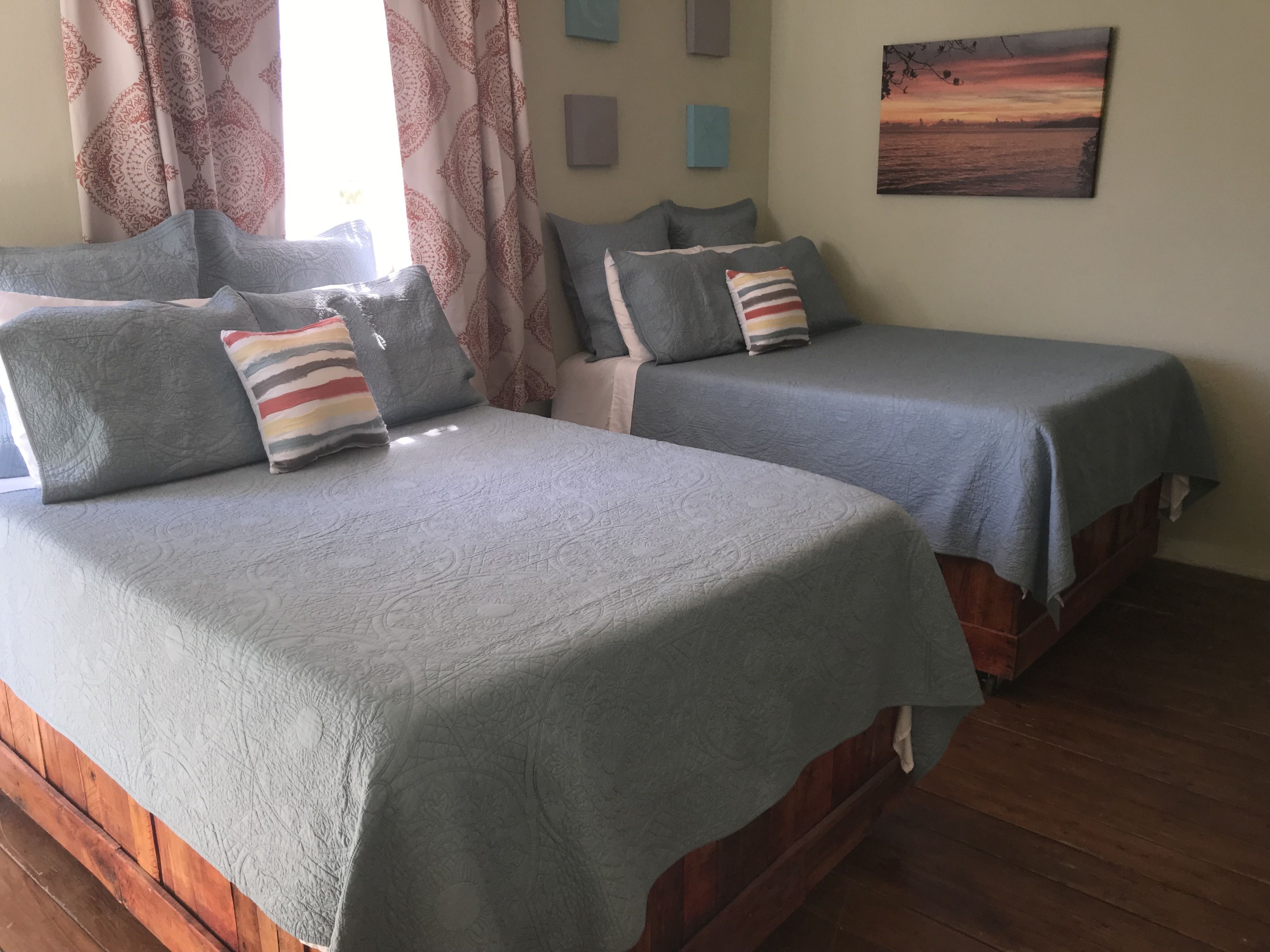 A room with two beds, each with a grey bedspread and pillows, a painting on the wall, and patterned curtains.