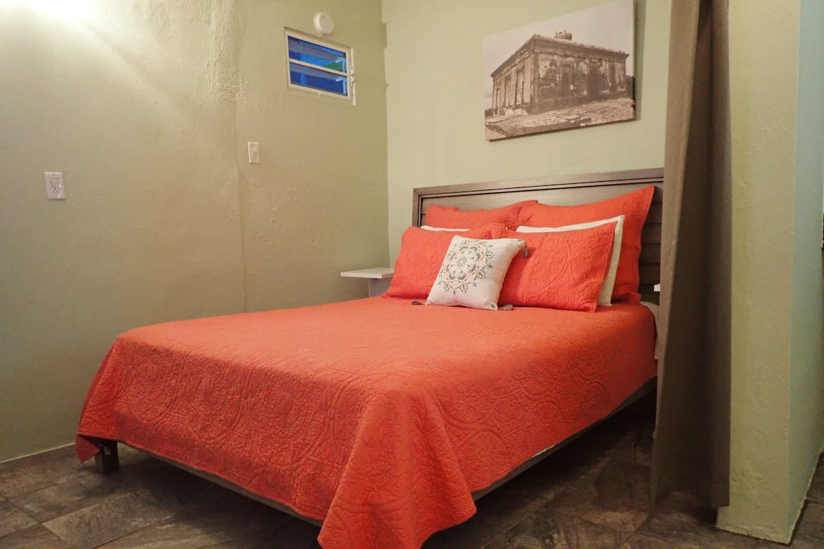 A single bed with a red bedspread and pillows, a small window, and a wall-mounted picture.