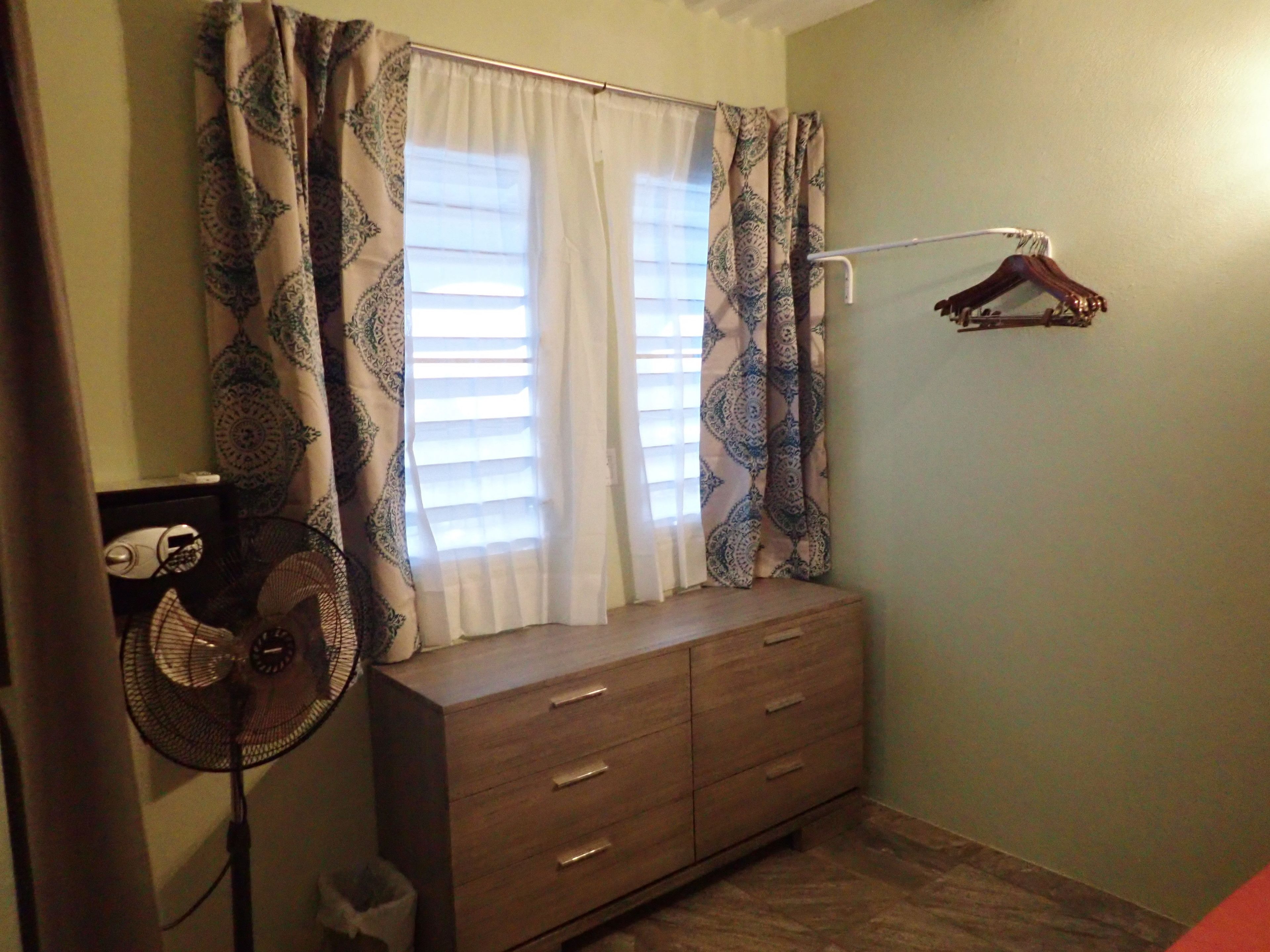 A bedroom with a wooden dresser, a hanging fan, patterned curtains, and a clothes hanger on the wall.