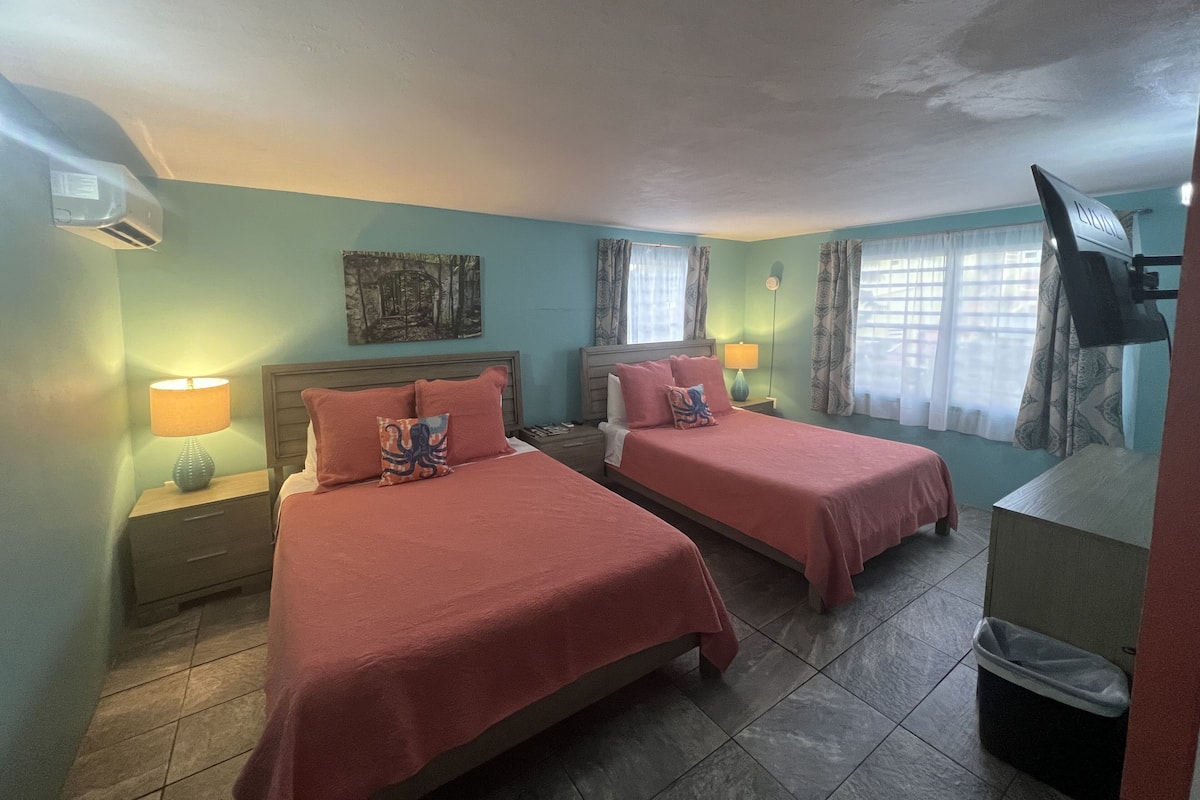 A hotel room with two beds, a nightstand with a lamp, a wall-mounted TV, and a ceiling fan.