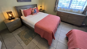 A bedroom with a bed, nightstands, a window with curtains, a painting on the wall, and a patterned rug. - The Vieques Guesthouse (Vieques)