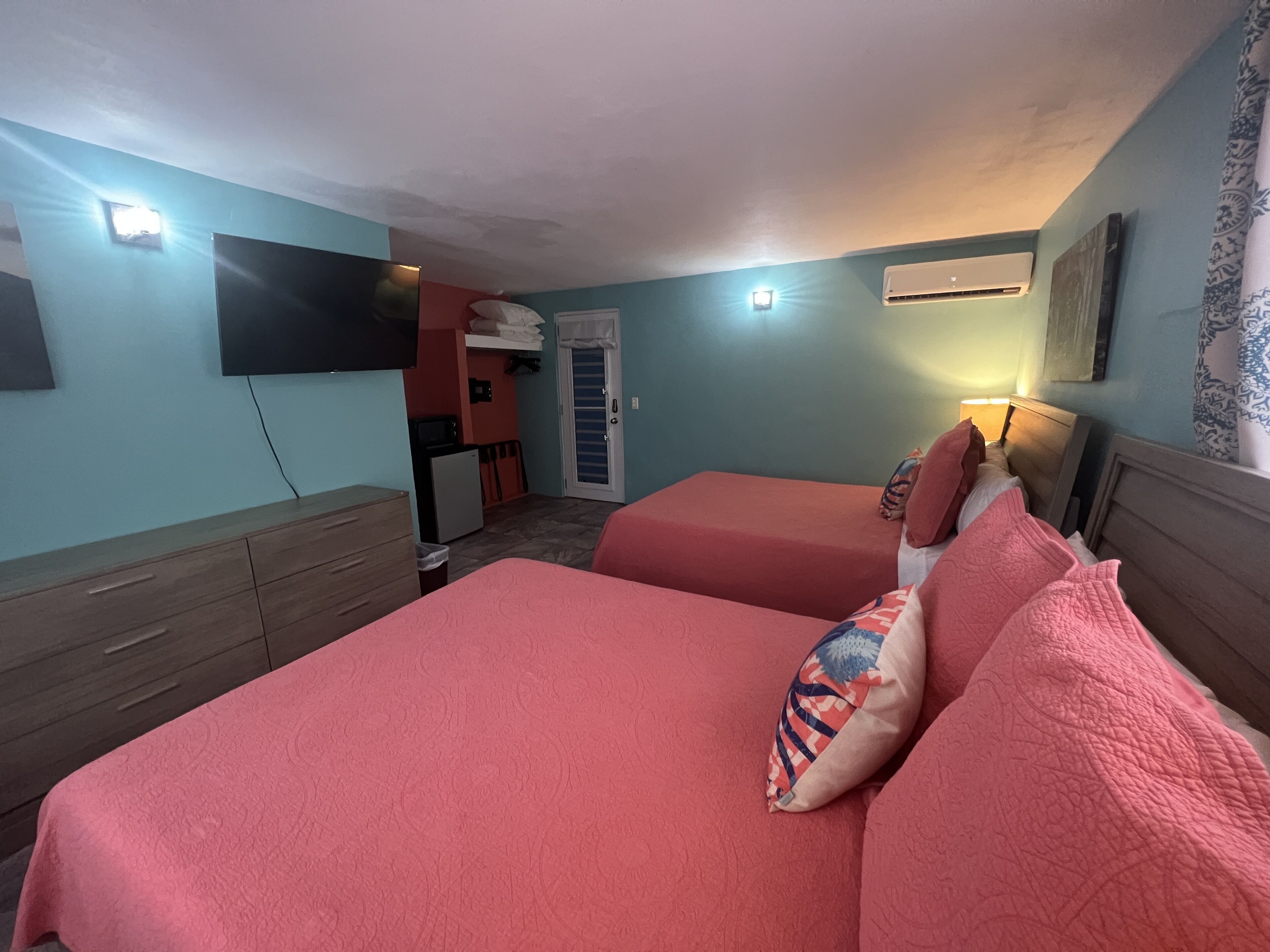 A room with two beds, a TV mounted on the wall, a small kitchenette, and an air conditioning unit.