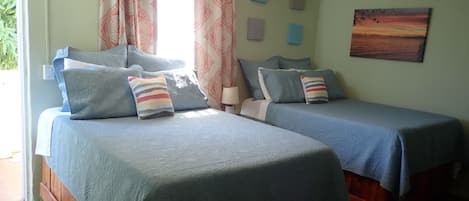 A room with two beds, each with a blue bedspread and multiple pillows. There is a ceiling fan, a window with patterned curtains, and a painting on the wall.
