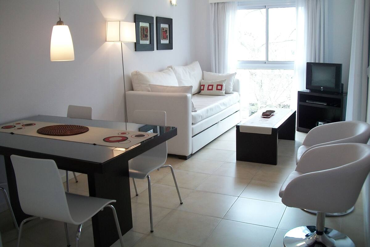 Una sala de estar moderna con un sofá blanco, una mesa de comedor con sillas, un mueble para la televisión y un ventilador de techo.