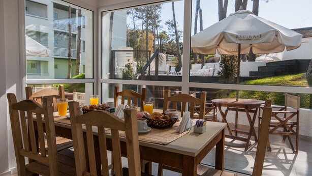 Una mesa de desayuno con sillas de madera, un paraguas blanco y vistas a una zona residencial a través de amplios ventanales.