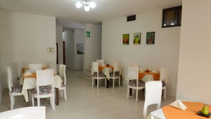 Un comedor con sillas blancas y manteles naranjas, una lámpara en el techo y cuadros de frutas colgados en la pared.