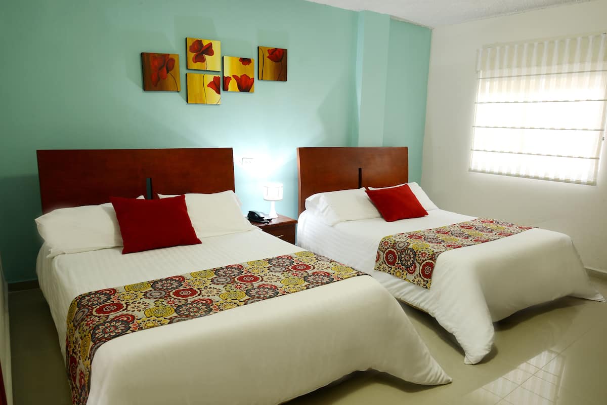 Habitación de hotel con dos camas, cada una con un almohadón rojo y una colcha estampada. Se observan obras de arte en las paredes sobre las camas y una ventana con persianas a la derecha.