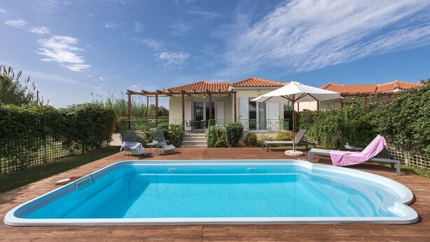 A swimming pool with a wooden deck, a covered patio with outdoor furniture, and a house with a tiled roof in the background. - Jewel of Venus Villa - Private Pool Paradise (Pylos-Nestoras)