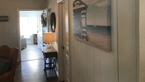 Um corredor com piso de madeira, uma porta, um abajur, uma cadeira de vime, uma pequena mesa com abajur e uma pintura de uma ponte sobre a água, com vista para outro quarto.