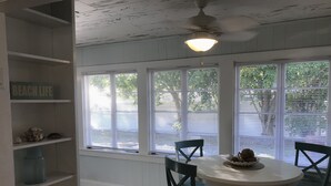 Uma sala de jantar com mesa redonda e quatro cadeiras, janelas amplas com persianas, ventilador de teto e uma prateleira com a placa 'BEACH LIFE'.