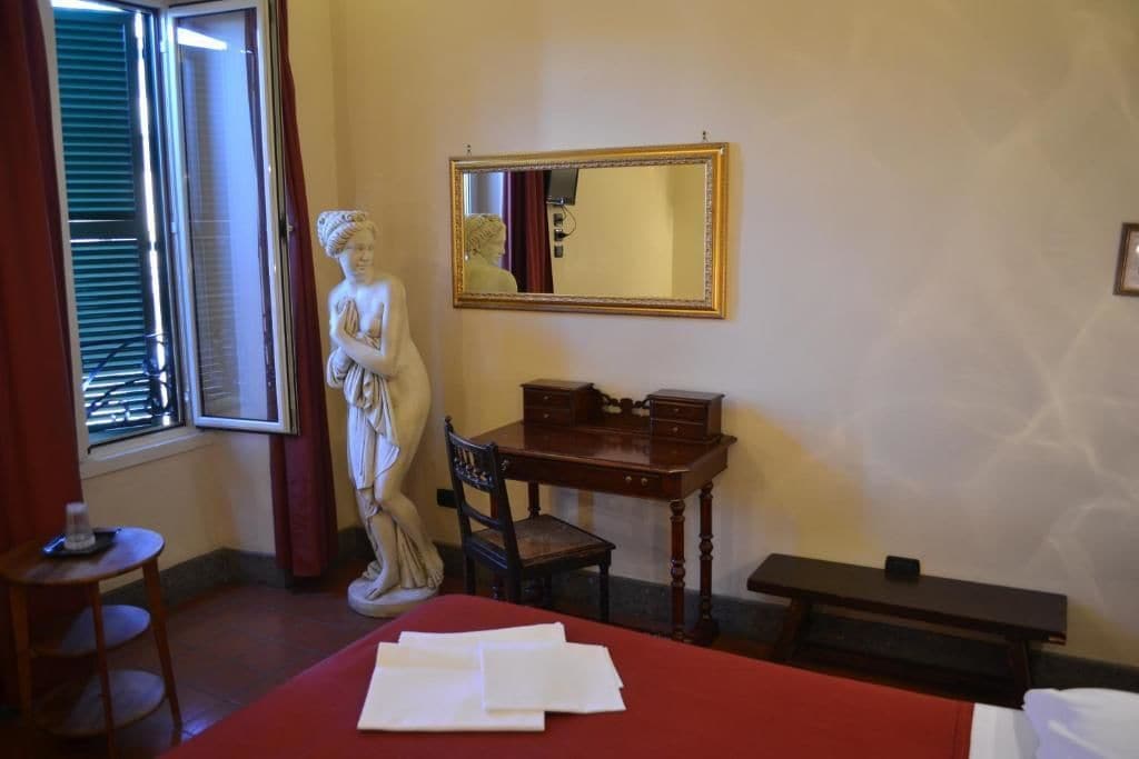 A room with a bed, a desk, a chair, a statue, a mirror, and a window with blinds.
