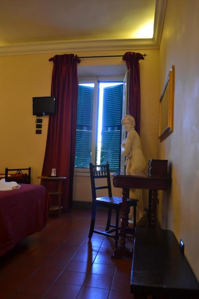 A room with a bed, a desk, a chair, a statue, a window with curtains, and a television.