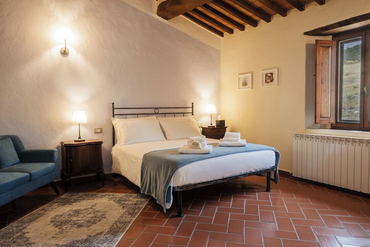 Une chambre à coucher avec un lit, des lampes de chevet, un fauteuil bleu, une table de chevet en bois et une fenêtre donnant sur un paysage verdoyant.