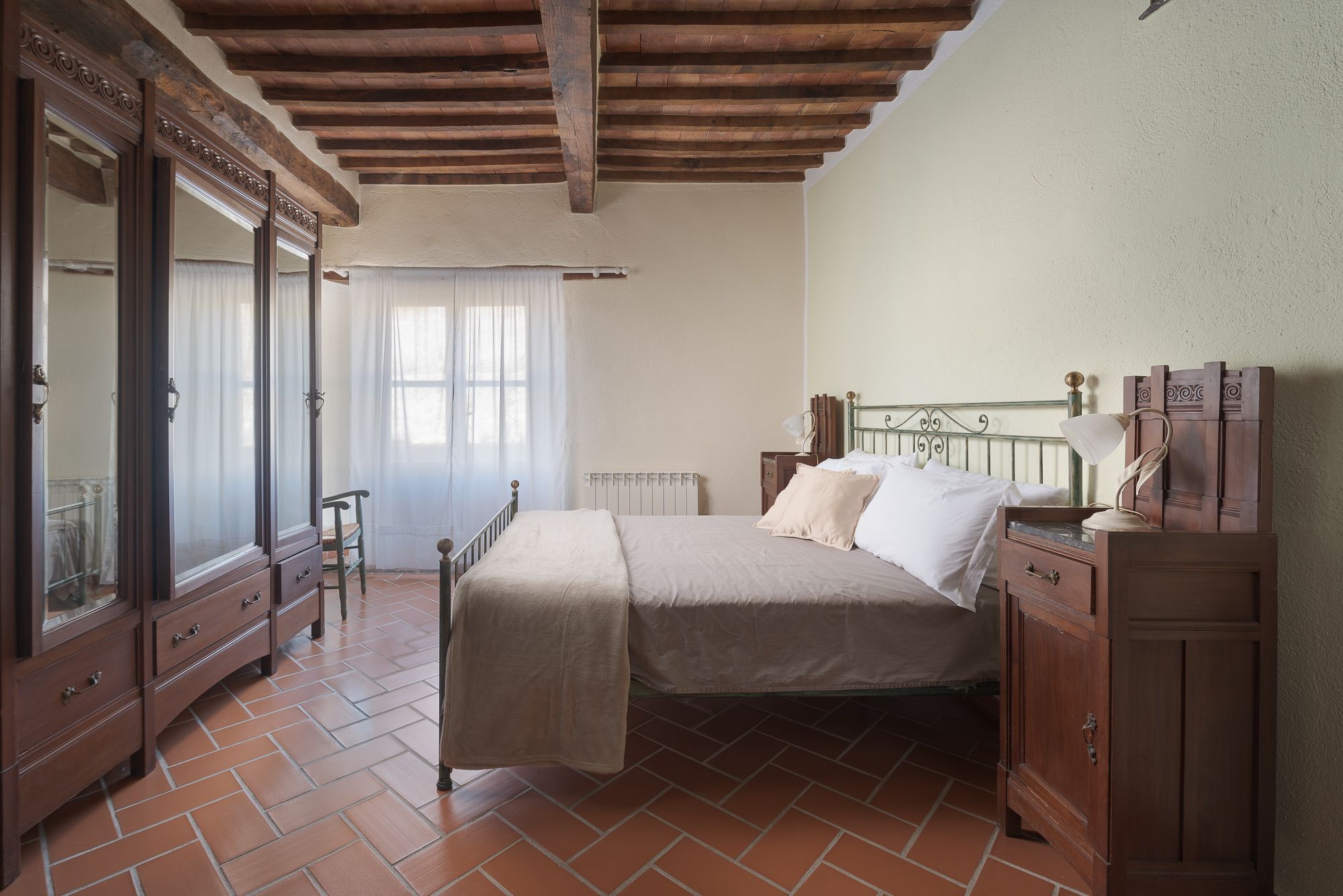 A bedroom with a bed, wooden furniture, a radiator, and a window with sheer curtains.