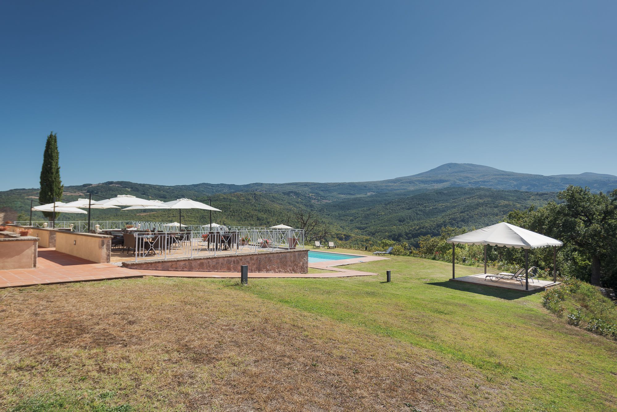 A pool area with outdoor seating and umbrellas, surrounded by a grassy landscape and hills in the background.