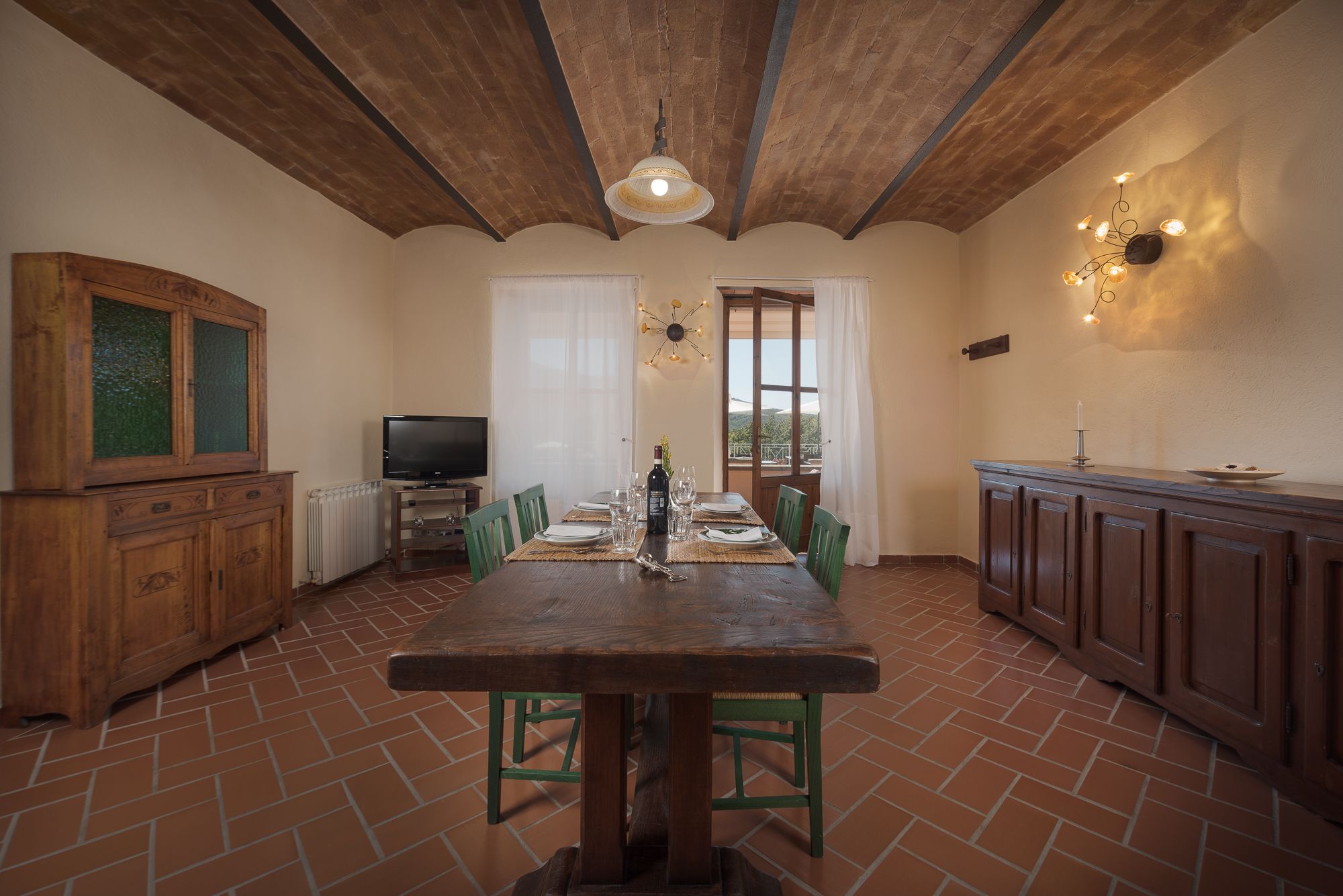 A dining area with a wooden table and chairs, a television, and a view to the outside.