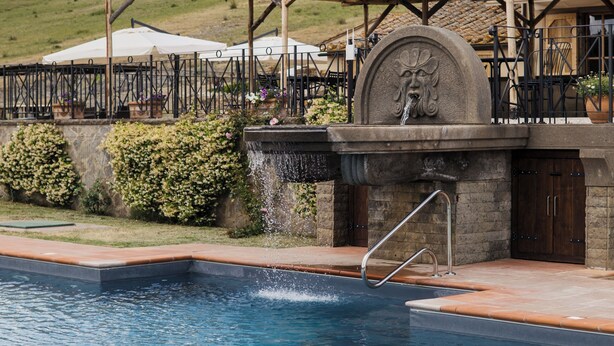 Una piscina con una fuente de piedra, rodeada por un patio cubierto con mobiliario exterior.